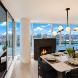 Dining room with a marble table set for six, large windows overlooking a river and cityscape, and a modern chandelier above a lit fireplace.