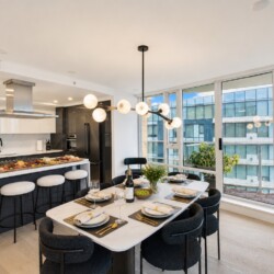 Open-plan kitchen and dining area with a marble table set for eight, gold cutlery, and a city skyline view through large windows.