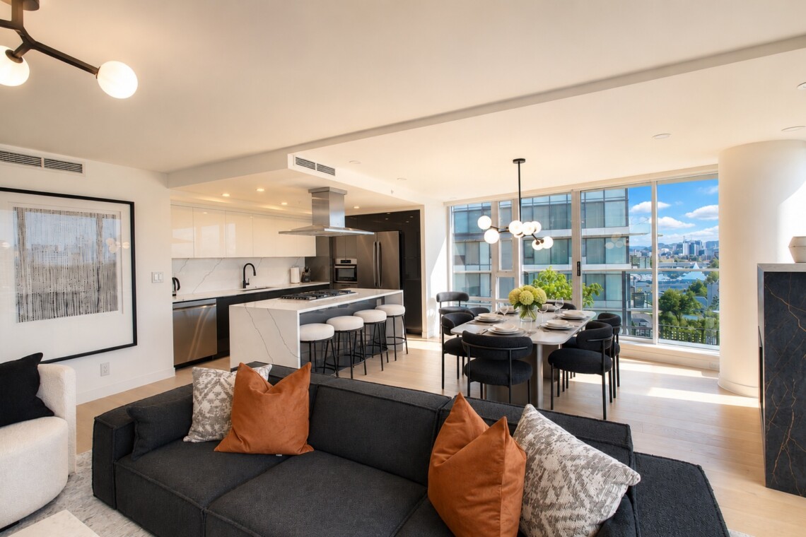 Open-plan living room and kitchen: dark sofa with orange cushions, marble island, and a dining table by large windows with a city view.
