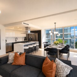 Open-plan living room and kitchen: dark sofa with orange cushions, marble island, and a dining table by large windows with a city view.