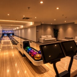 Indoor bowling alley with polished wooden lanes, a ball return displaying colorful bowling balls, and a seating area with white couches in the background.