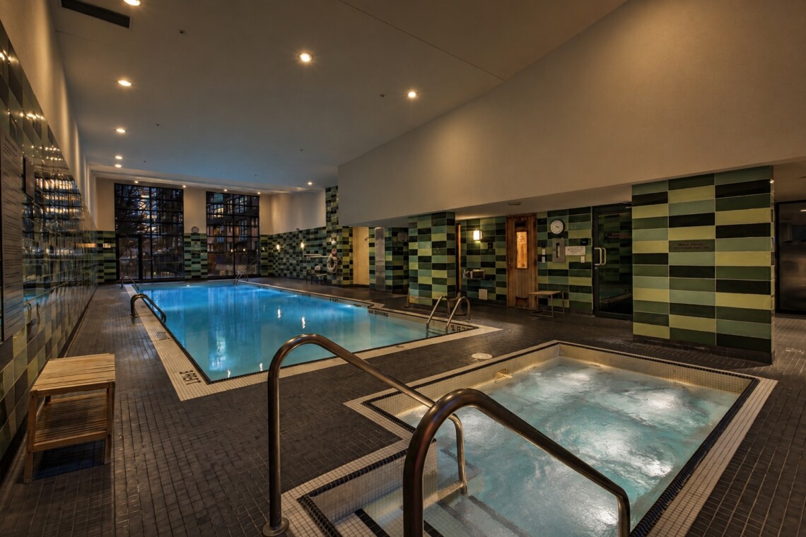 Indoor swimming pool area in a spa with green tiled walls, metal handrails, and a connected hot tub in the foreground.