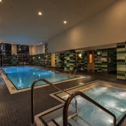 Indoor swimming pool area in a spa with green tiled walls, metal handrails, and a connected hot tub in the foreground.