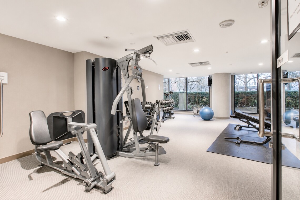 Modern gym with a multi-station weight machine, benches, dumbbells, and a blue exercise ball near large windows.