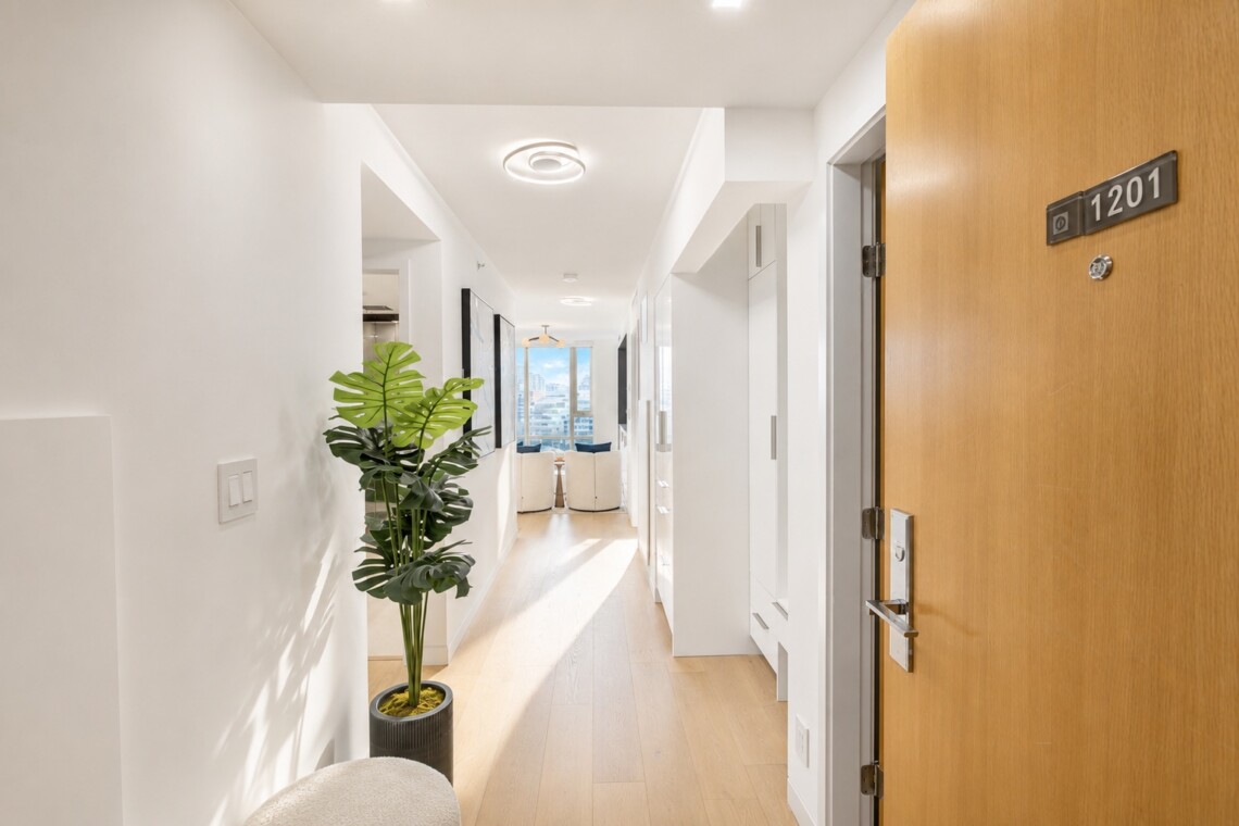 Bright white hallway with light wood floor, a tall potted plant, and a living room with a city view at the end.