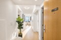 Bright white hallway with light wood floor, a tall potted plant, and a living room with a city view at the end.