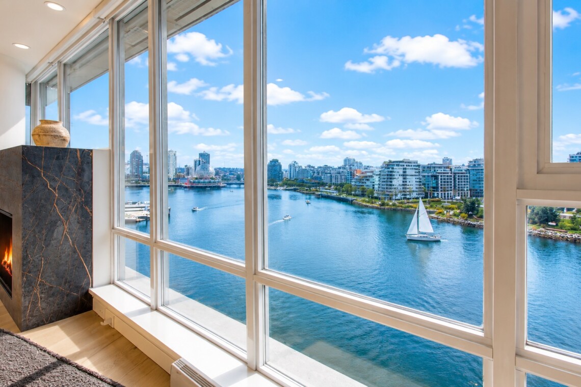 Living room with a marble fireplace and large windows overlooking a river, sailboat, and city skyline.