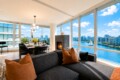 Bright open living-dining space with floor-to-ceiling windows showing a river and city skyline, a marble fireplace, and a black sectional with orange cushions?