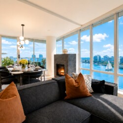 Bright open living-dining space with floor-to-ceiling windows showing a river and city skyline, a marble fireplace, and a black sectional with orange cushions?