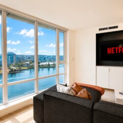 Modern living room with oversized windows offering a river and city view; Netflix is displayed on the wall TV.