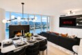 Open-plan living and dining area with a lit fireplace, marble dining table set for six, and floor-to-ceiling windows overlooking a river cityscape.