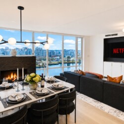Open-plan living and dining area with a lit fireplace, marble dining table set for six, and floor-to-ceiling windows overlooking a river cityscape.