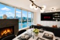 Bright, modern living and dining area with floor-to-ceiling windows overlooking a river and city skyline; marble table set for six with candles and flowers, and a Netflix screen on the wall.