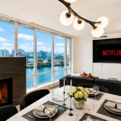 Bright, modern living and dining area with floor-to-ceiling windows overlooking a river and city skyline; marble table set for six with candles and flowers, and a Netflix screen on the wall.