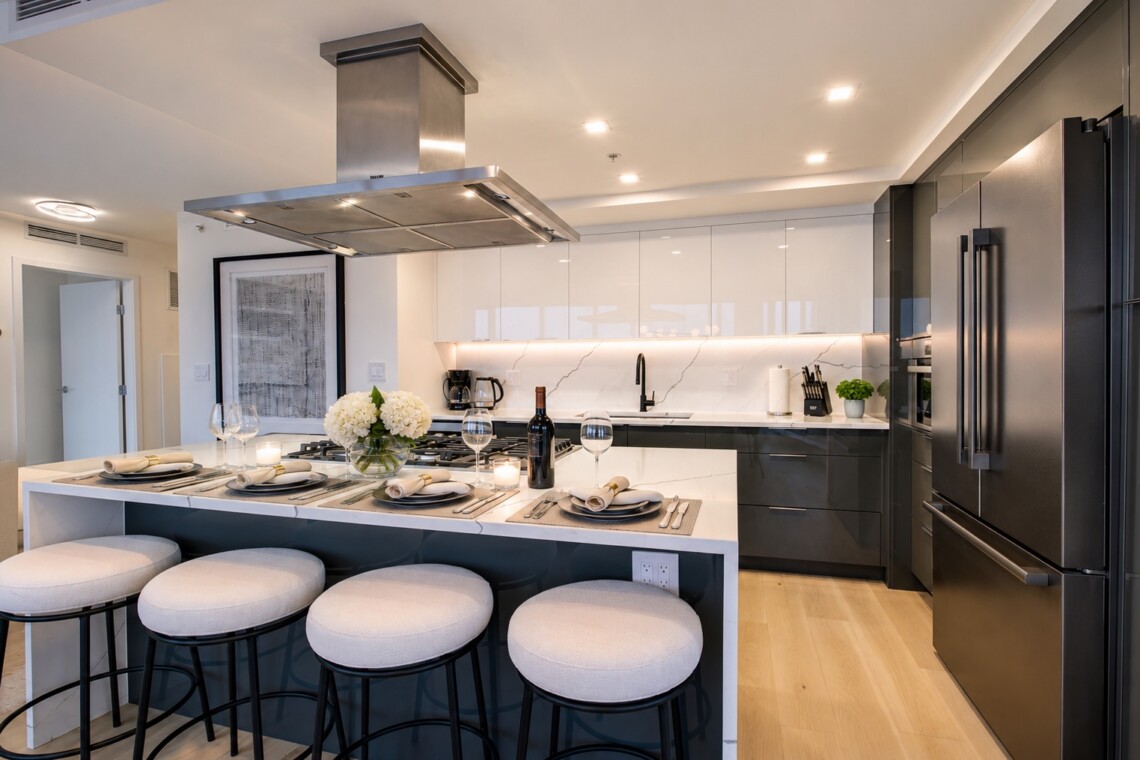 Modern kitchen with a marble island, white and dark cabinets, and four cushioned stools set for a meal with wine and flowers on the table.