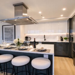 Modern kitchen with a marble island, white and dark cabinets, and four cushioned stools set for a meal with wine and flowers on the table.