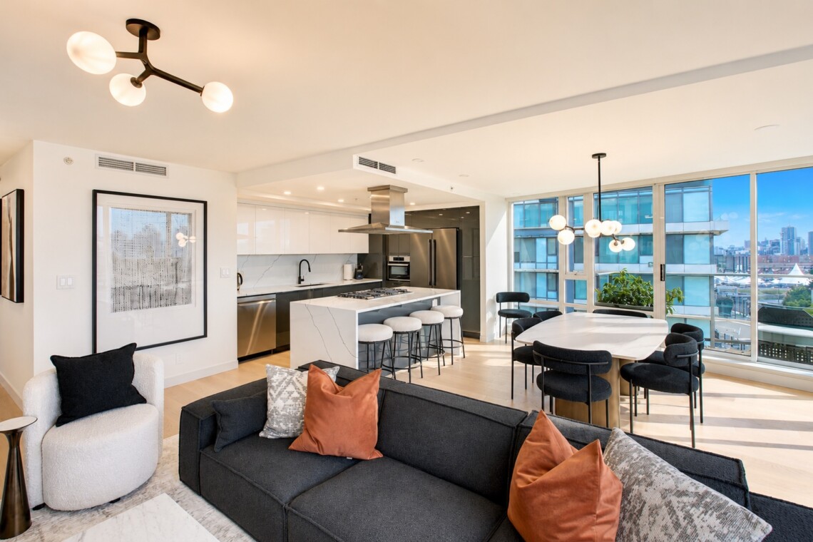 Open-concept living area with a white marble kitchen island, black sofa, and large windows overlooking a cityscape.