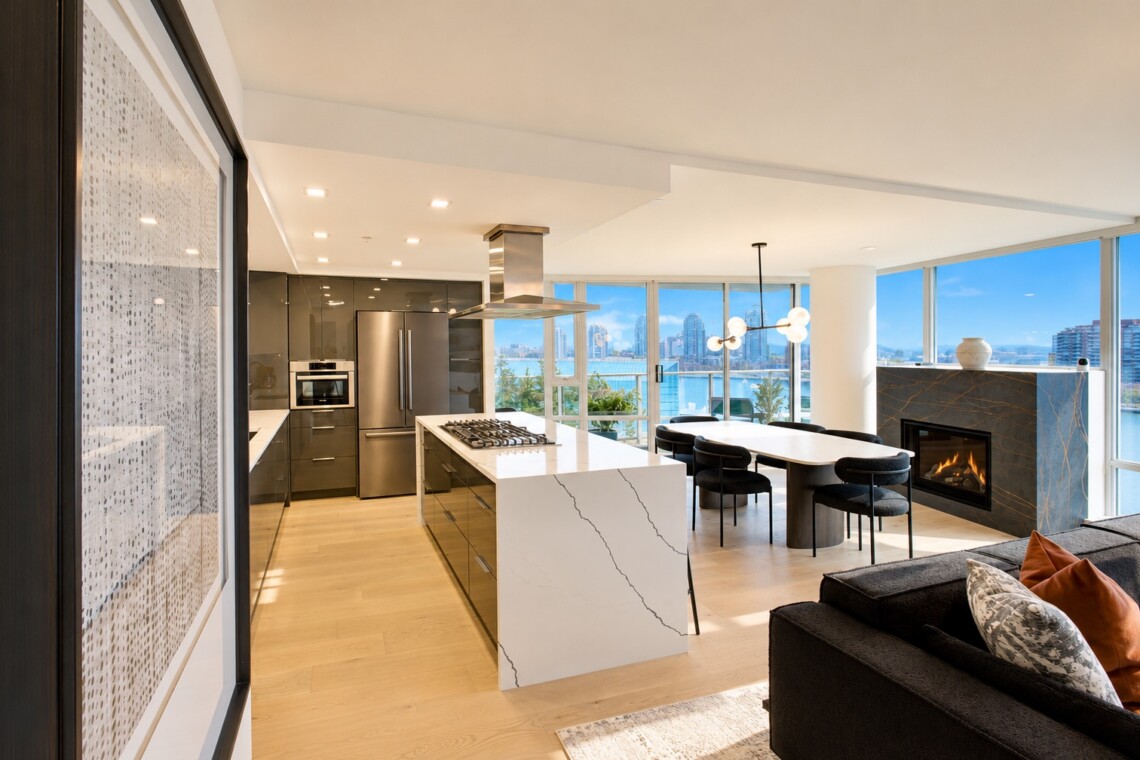 Open-concept kitchen with white marble island, stainless steel appliances, and a dining area by large windows overlooking water and city skyline.