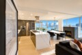 Open-concept kitchen with white marble island, stainless steel appliances, and a dining area by large windows overlooking water and city skyline.