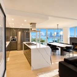 Open-concept kitchen with white marble island, stainless steel appliances, and a dining area by large windows overlooking water and city skyline.