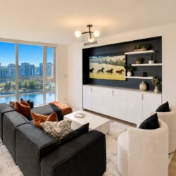 Modern living room with a dark gray sectional, orange pillows, and white chairs, overlooking a river and city skyline through floor-to-ceiling windows.