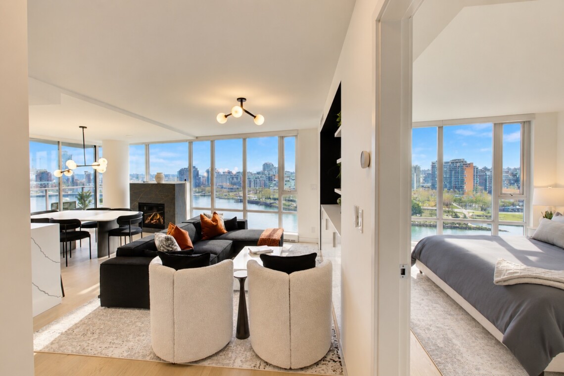 Bright, open-concept living space with large windows overlooking a river and city skyline; contemporary furniture, fireplace, and dining area, plus a adjacent bedroom visible through a doorway.