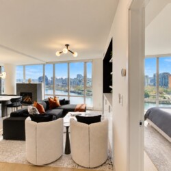 Bright, open-concept living space with large windows overlooking a river and city skyline; contemporary furniture, fireplace, and dining area, plus a adjacent bedroom visible through a doorway.
