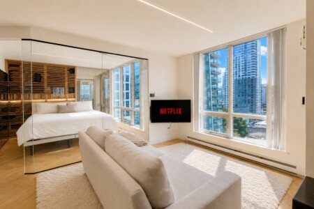 Modern, minimalist studio with a glass partition, white bedding, beige sofa, and a wall-mounted Netflix screen, city skyline outside the windows.