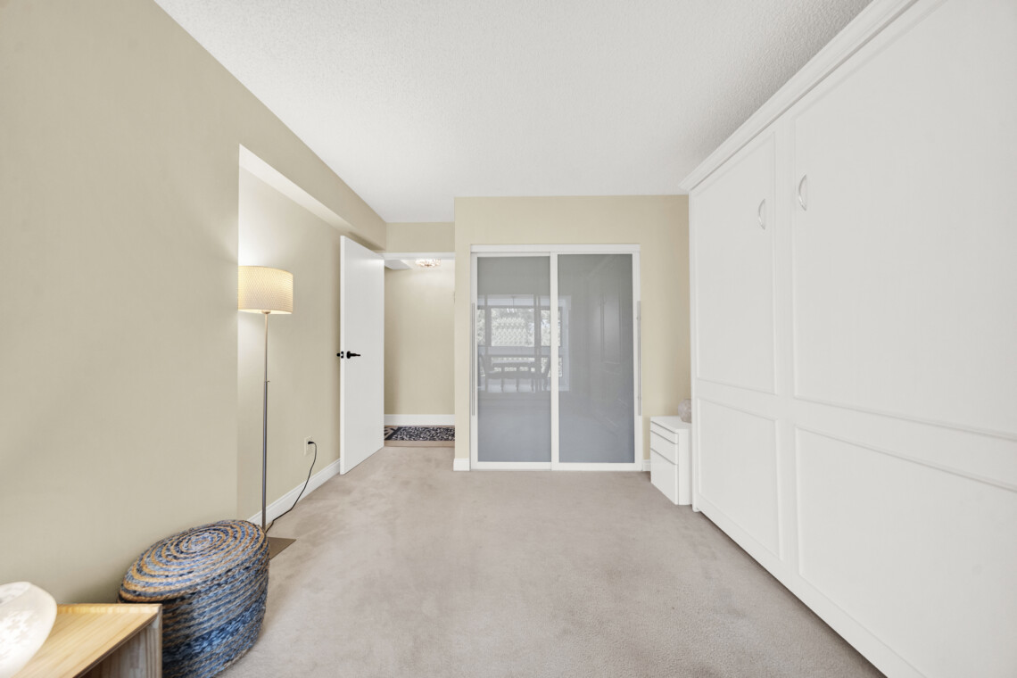 Bright beige hallway with white built-in cabinets on the right and a sliding glass door at the end; a floor lamp and woven ottoman sit on the left.