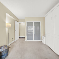 Bright beige hallway with white built-in cabinets on the right and a sliding glass door at the end; a floor lamp and woven ottoman sit on the left.