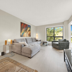 Bright living room with a gray sectional sofa, glass coffee table, abstract wall art, and large windows overlooking trees outside.