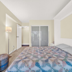Bedroom with pastel yellow walls, a bed covered by a colorful zigzag quilt and a gray pillow, wicker pouf, and a floor lamp on the left.