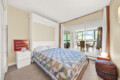 Bedroom with a white bed frame and a colorful patchwork quilt, white nightstands, and mirrored closet doors reflecting a sunny living area outside the room.