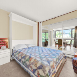Bedroom with a white bed frame and a colorful patchwork quilt, white nightstands, and mirrored closet doors reflecting a sunny living area outside the room.