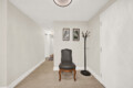 Narrow hallway with beige walls, a black upholstered chair, and a tall coat rack beside two framed landscape prints on the wall.