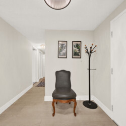 Narrow hallway with beige walls, a black upholstered chair, and a tall coat rack beside two framed landscape prints on the wall.