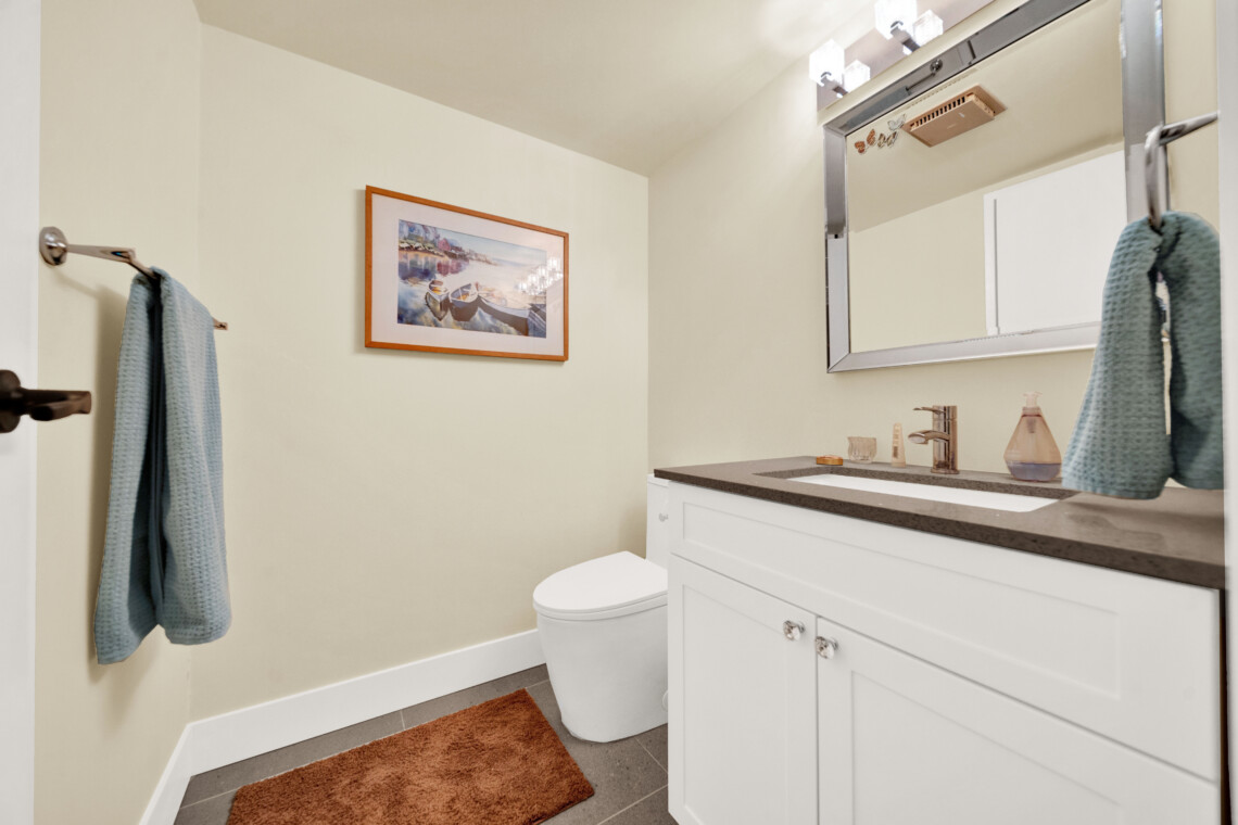 Small bathroom with a white vanity, gray countertop, framed harbor scene, and blue towels hanging nearby; toilet in the corner.