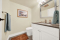 Small bathroom with a white vanity, gray countertop, framed harbor scene, and blue towels hanging nearby; toilet in the corner.