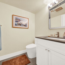 Small bathroom with a white vanity, gray countertop, framed harbor scene, and blue towels hanging nearby; toilet in the corner.