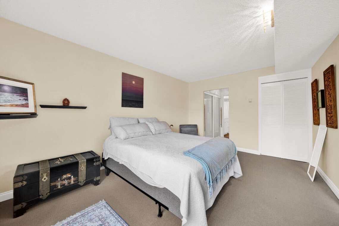 Bedroom with a queen bed, white quilt, blue throw, and two black decorative trunks at the foot of the bed on a beige wall.