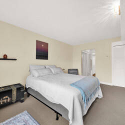 Bedroom with a queen bed, white quilt, blue throw, and two black decorative trunks at the foot of the bed on a beige wall.