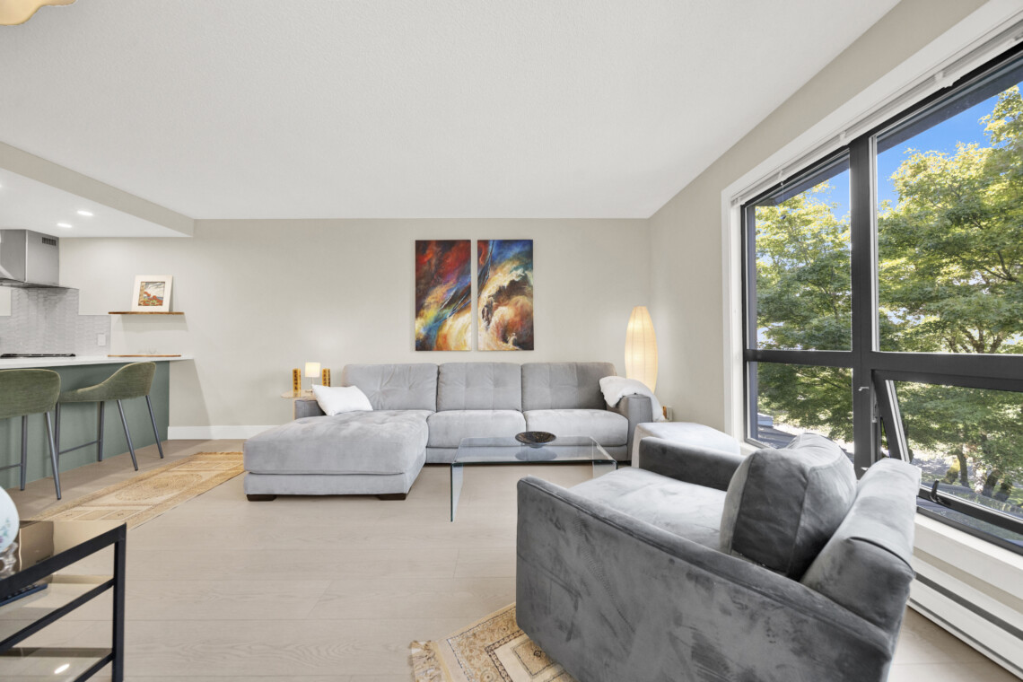 Bright living room with a light gray sectional sofa, glass coffee table, and two-panel abstract paintings on the wall near a large window view of trees.