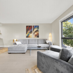 Bright living room with a light gray sectional sofa, glass coffee table, and two-panel abstract paintings on the wall near a large window view of trees.