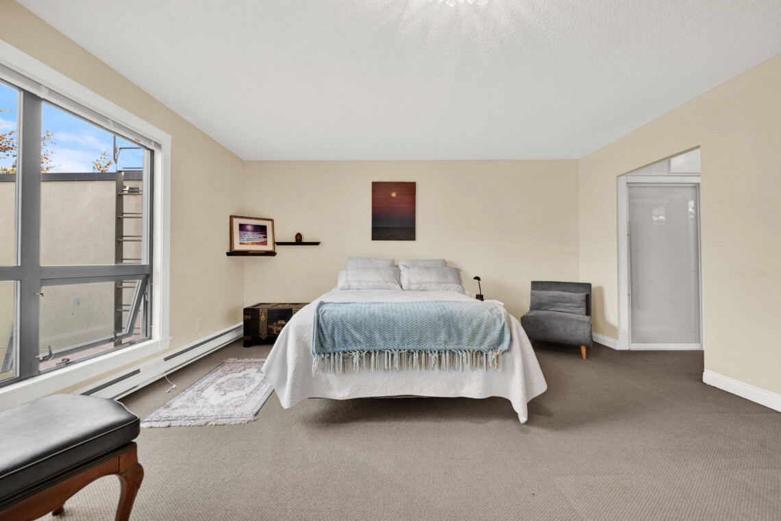 Bedroom with a queen bed centered, light blue blanket, beige walls, and a gray armchair by the right wall; large window on the left fills the room with daylight.