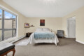 Bedroom with a queen bed centered, light blue blanket, beige walls, and a gray armchair by the right wall; large window on the left fills the room with daylight.