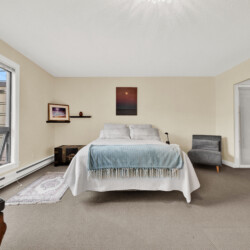 Bedroom with a queen bed centered, light blue blanket, beige walls, and a gray armchair by the right wall; large window on the left fills the room with daylight.