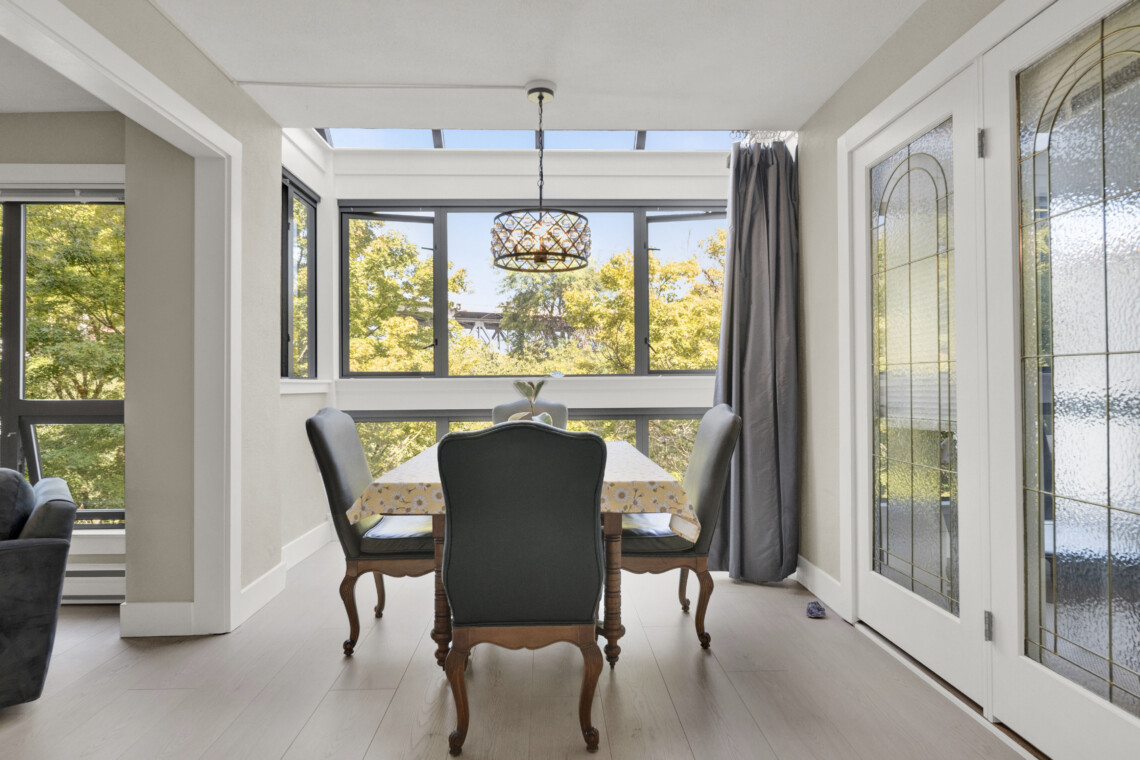 Sunlit dining area with a wooden table and floral tablecloth, carved chairs, and large windows offering an outdoor view; pendant light hangs above.”