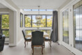 Sunlit dining area with a wooden table and floral tablecloth, carved chairs, and large windows offering an outdoor view; pendant light hangs above.”