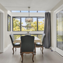 Sunlit dining area with a wooden table and floral tablecloth, carved chairs, and large windows offering an outdoor view; pendant light hangs above.”