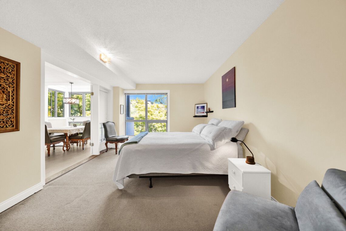 Bedroom with white bedding and beige walls; a nightstand sits beside the bed, with a view into a bright dining area through large windows.
